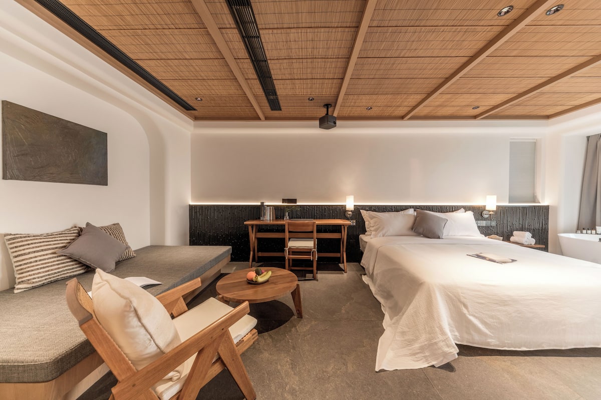 A hotel room with a bed, a sofa, a wooden table with fruit, a desk, and a chair.