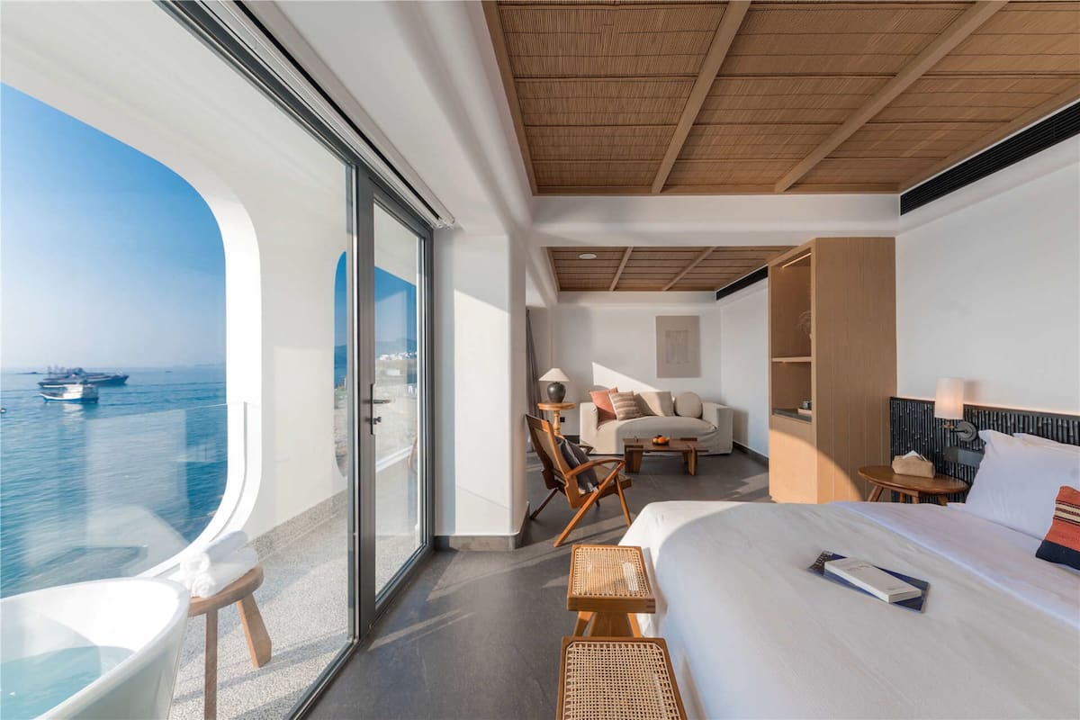 A modern hotel room with a large window overlooking the sea, a bed, a seating area with a table, and a small bookshelf.
