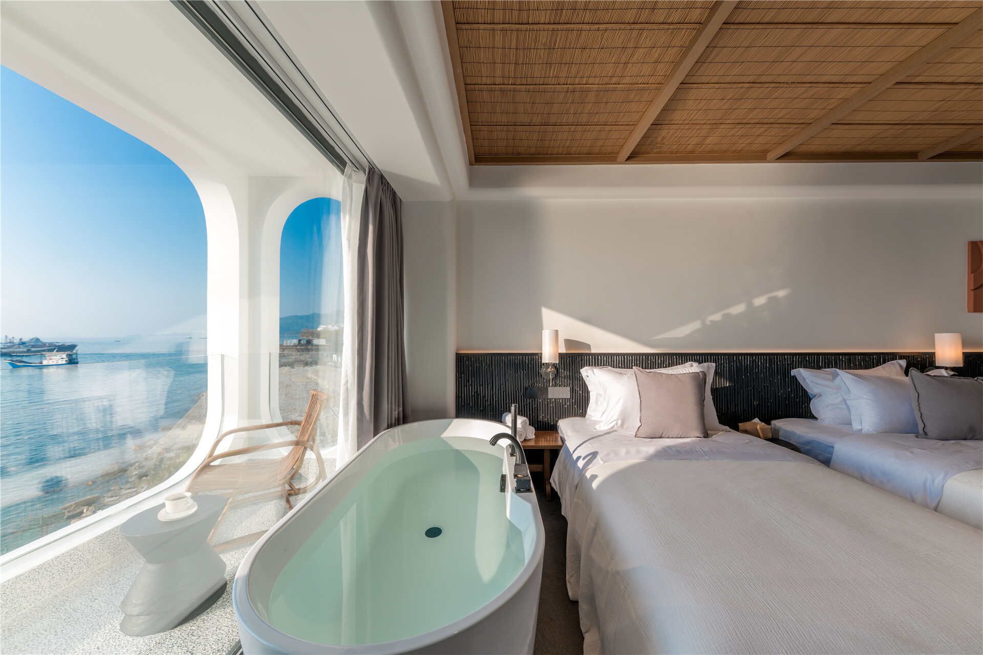 A modern hotel room with a large window offering a sea view, a bathtub, a bed with white bedding, and a wooden ceiling.