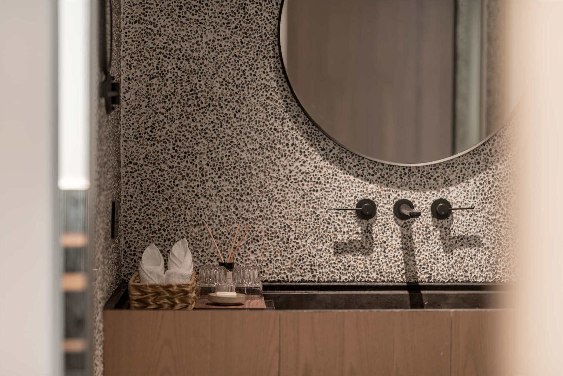A bathroom with a speckled tile backsplash, a round mirror, and a wooden vanity with a sink and glassware.