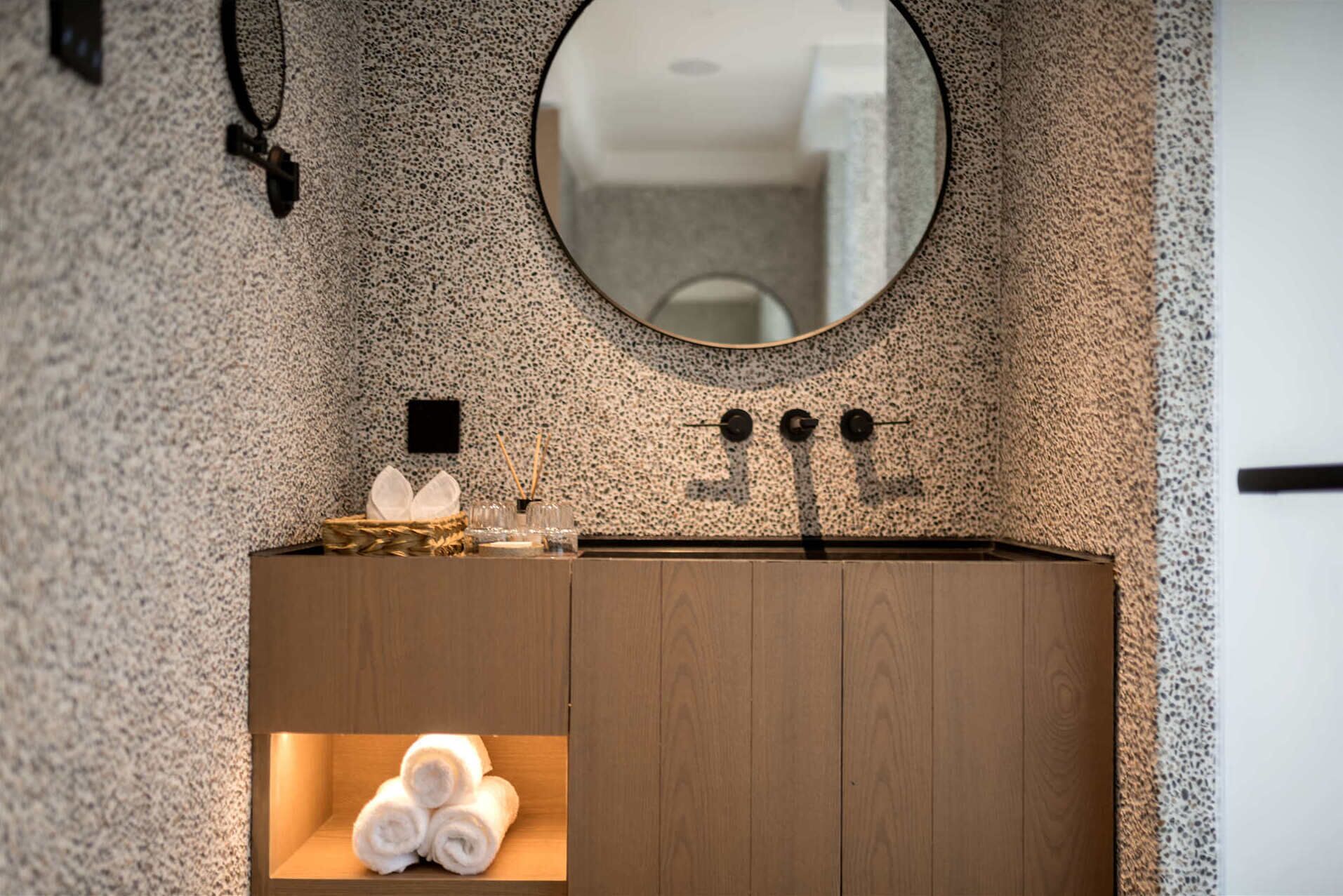 Un bagno con parete decorata, uno specchio rotondo, un mobile bagno in legno e uno scaffale con asciugamani arrotolati.