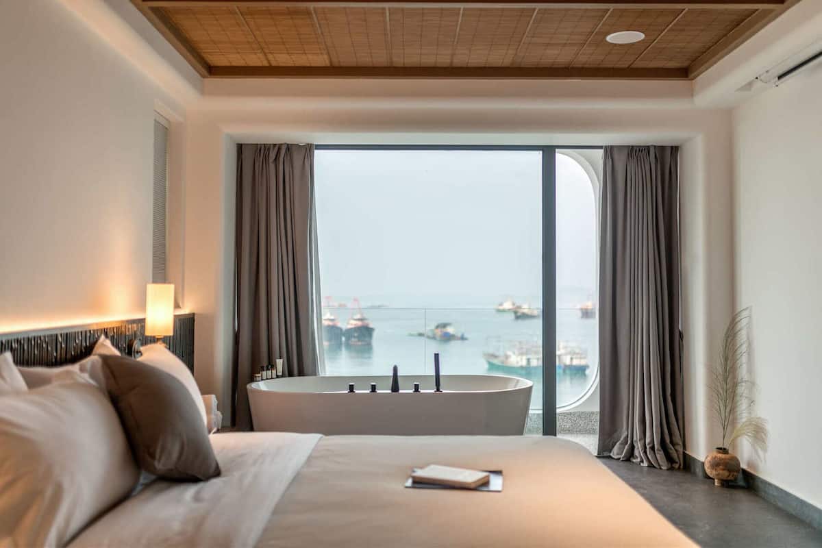 A modern hotel room with a large bed, a round bathtub, and a view of boats on the water.