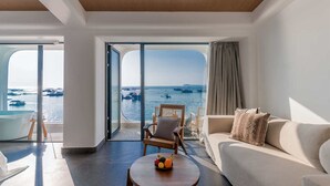 Un salón moderno con un gran ventanal corredero que da a un balcón con vistas al mar y barcos. Hay un sofá, una silla y una pequeña mesa con fruta encima.