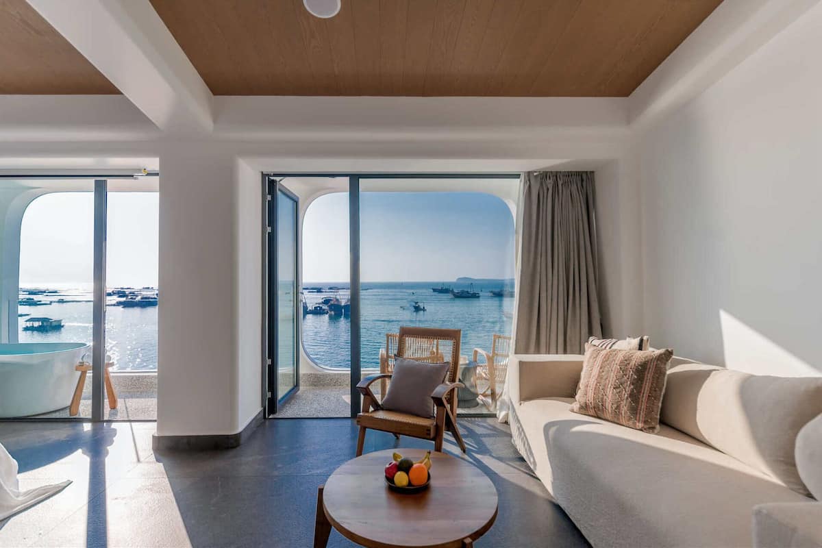 A modern living room with a large sliding glass door leading to a balcony with a view of the sea and boats. There is a sofa, a chair, and a small table with fruit on it.