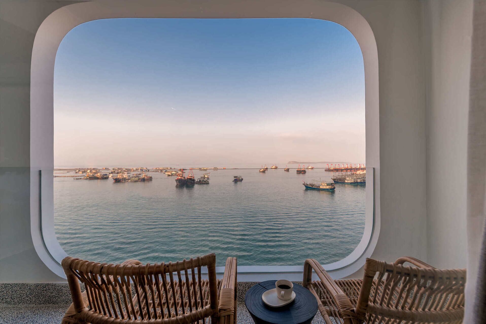 A view from a window with wicker chairs and a table, overlooking a calm sea with boats and a distant bridge.