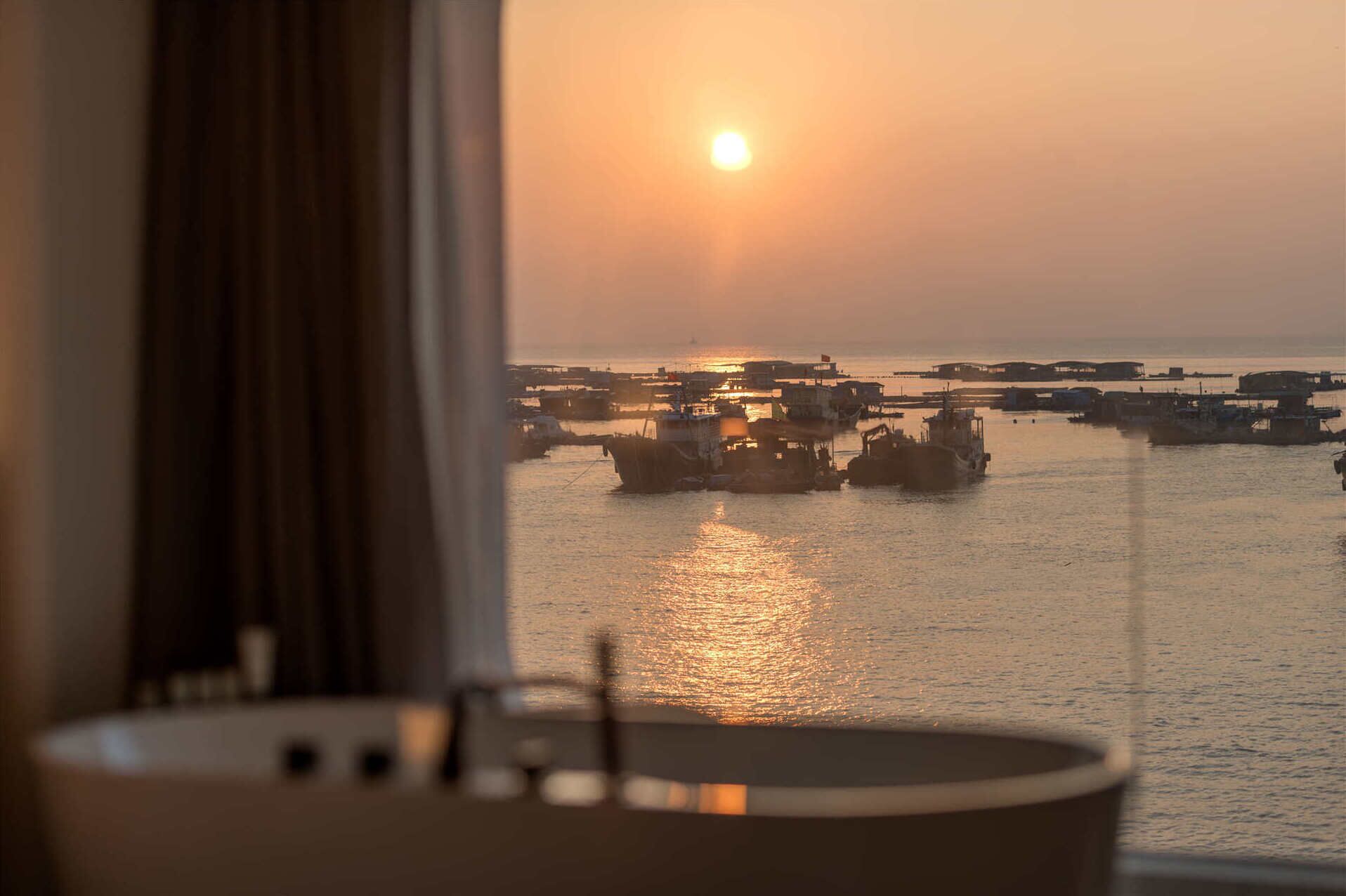 A view of a harbor with boats at sunset, seen through a window with a curtain.