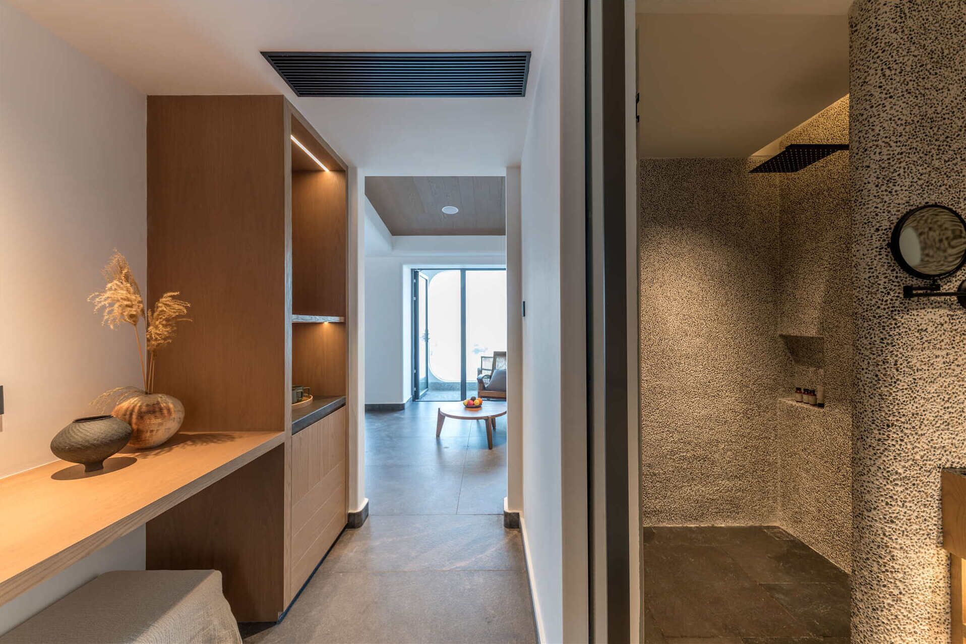 A modern interior with a wooden bench, a stone wall, and a view into a bright room with a table and chairs.
