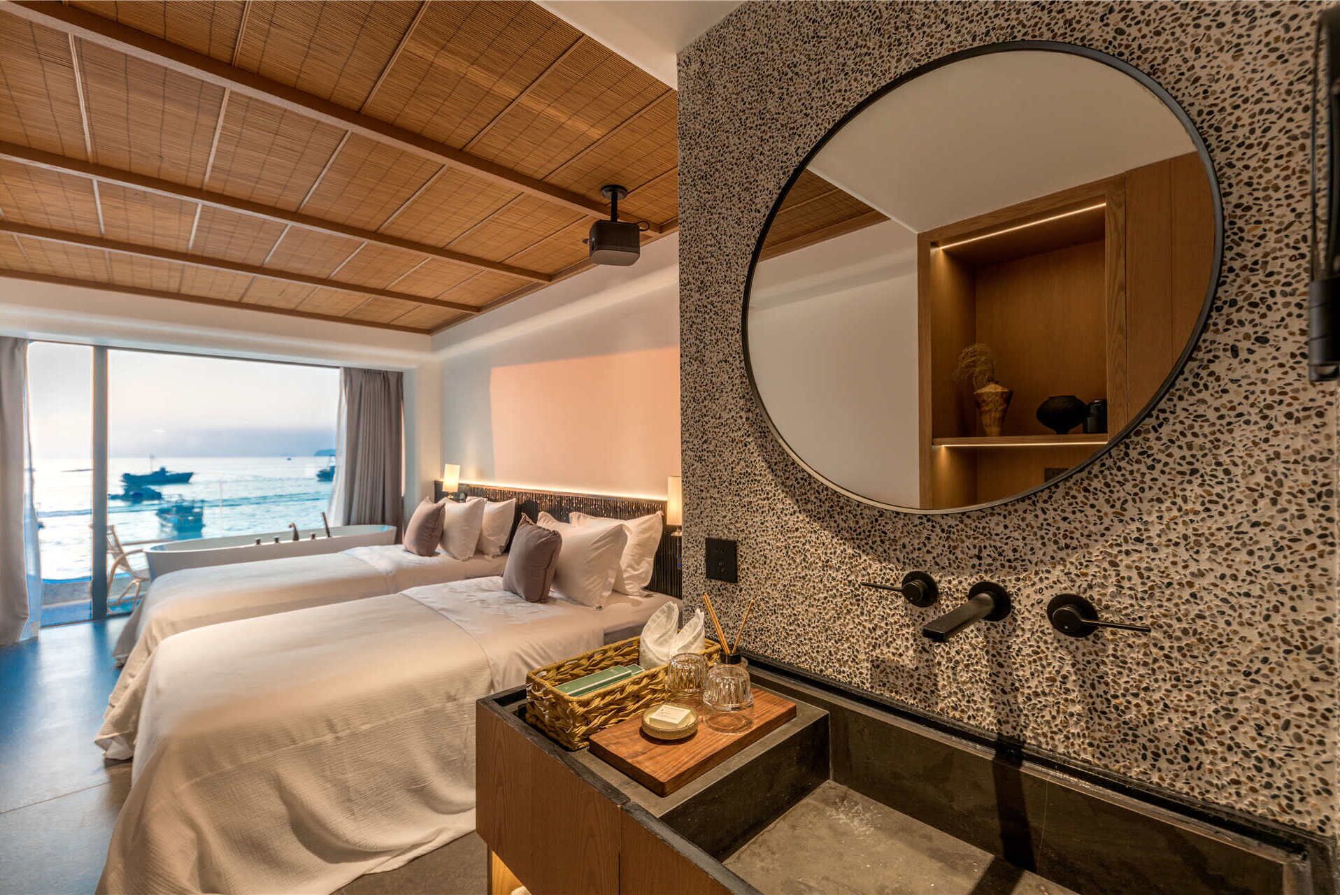 A hotel room with a large bed, a round mirror, a wooden headboard, a nightstand with a lamp, and a view of the sea through a large window.