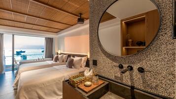 A hotel room with a large bed, a round mirror, a wooden headboard, a nightstand with a lamp, and a view of the sea through a large window.