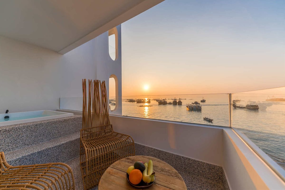 A balcony with a view of the sea at sunset, a wicker chair, a small table with fruit, and a bathtub.