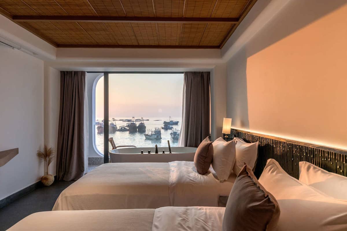 A hotel room with two beds, a large window with a view of boats, a wooden ceiling, and a modern headboard.