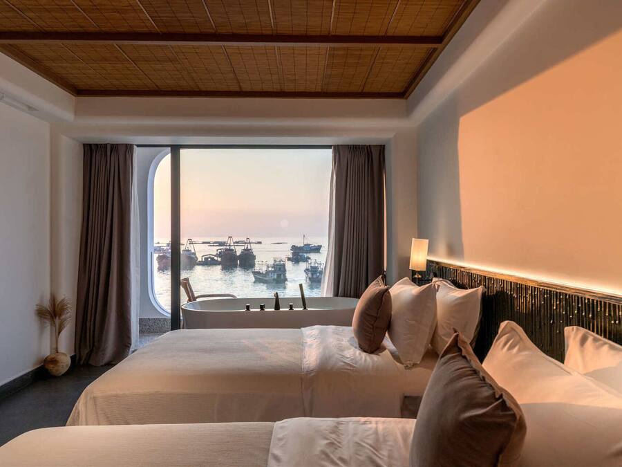 A hotel room with two beds, a large window with a view of boats, a wooden ceiling, and a modern headboard.