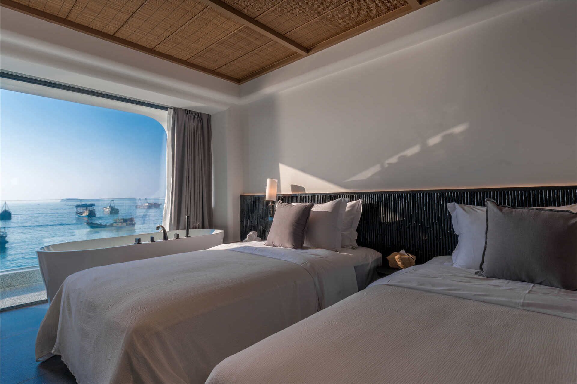A hotel room with two beds, a large window with a view of the sea, and a ceiling with a wooden panel design.