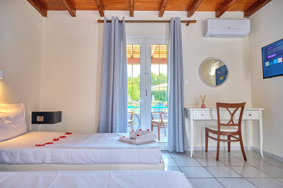 A hotel room with two beds, a desk, a chair, a round mirror, and a view of the outside.