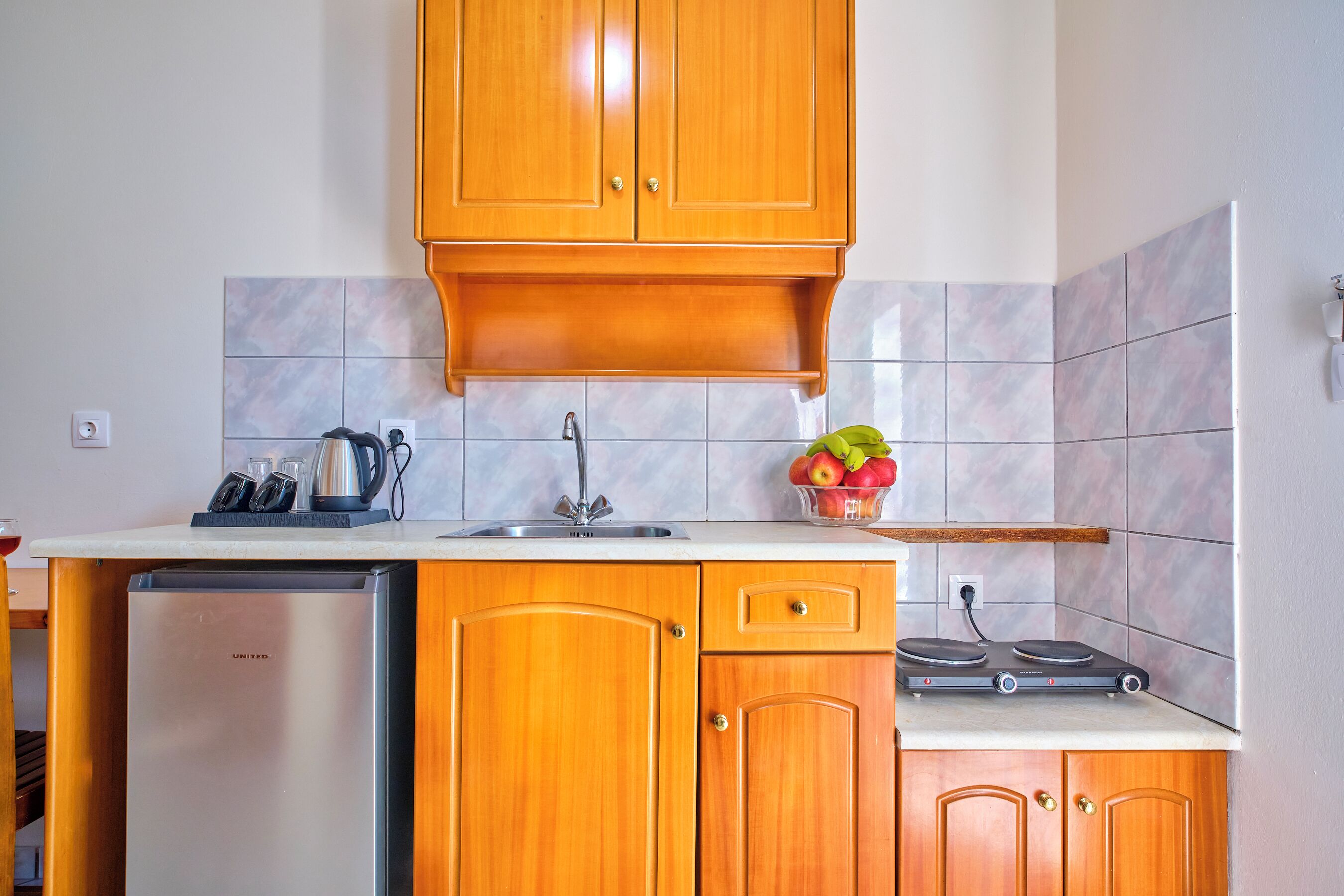A compact kitchen with wooden cabinets, a stainless steel refrigerator, a sink, and a small countertop with a kettle, glasses, and a fruit bowl.