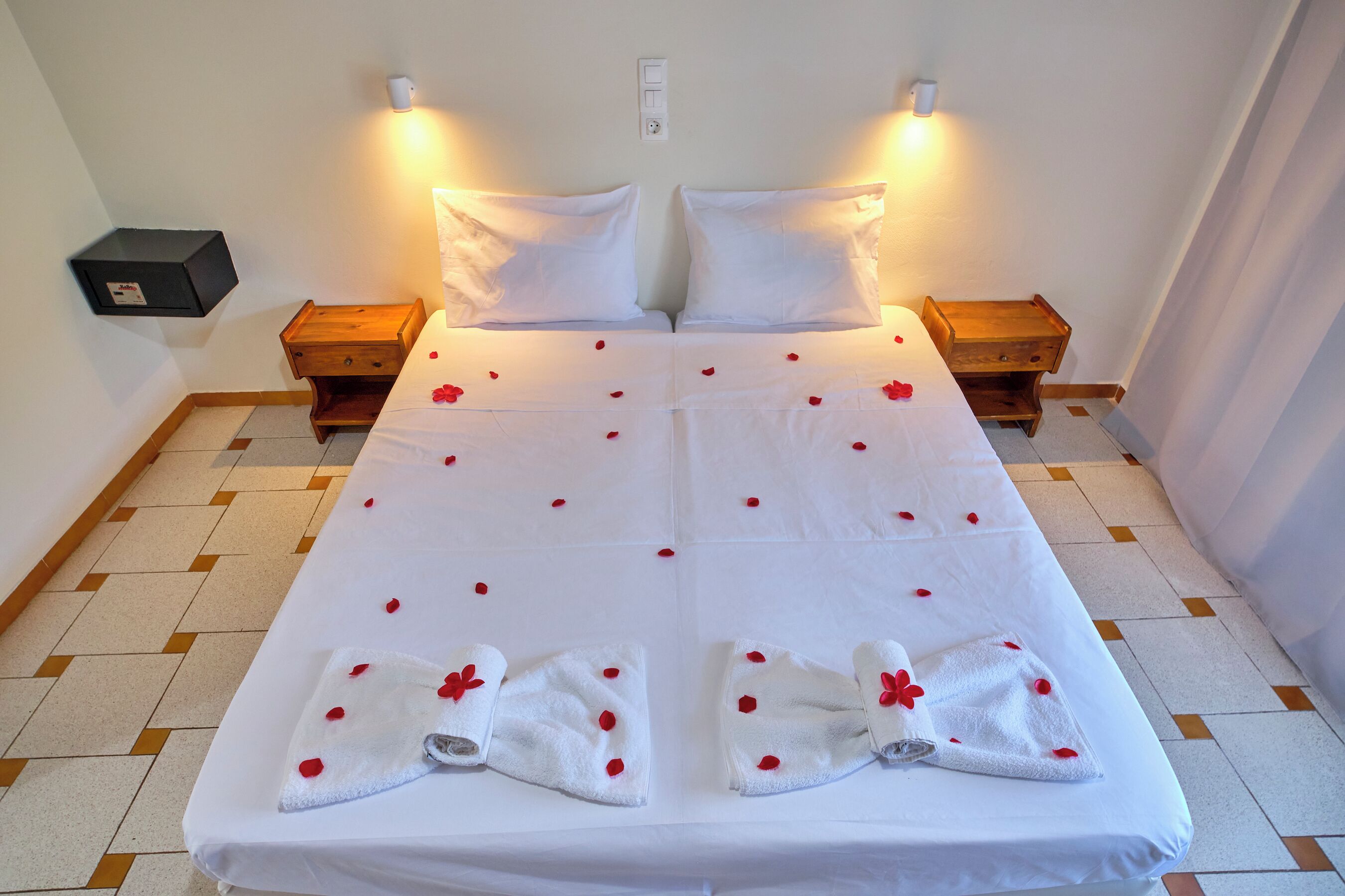 Un lit double décoré de pétales de rose et agrémenté de serviettes, deux tables de chevet et un luminaire fixé au mur.