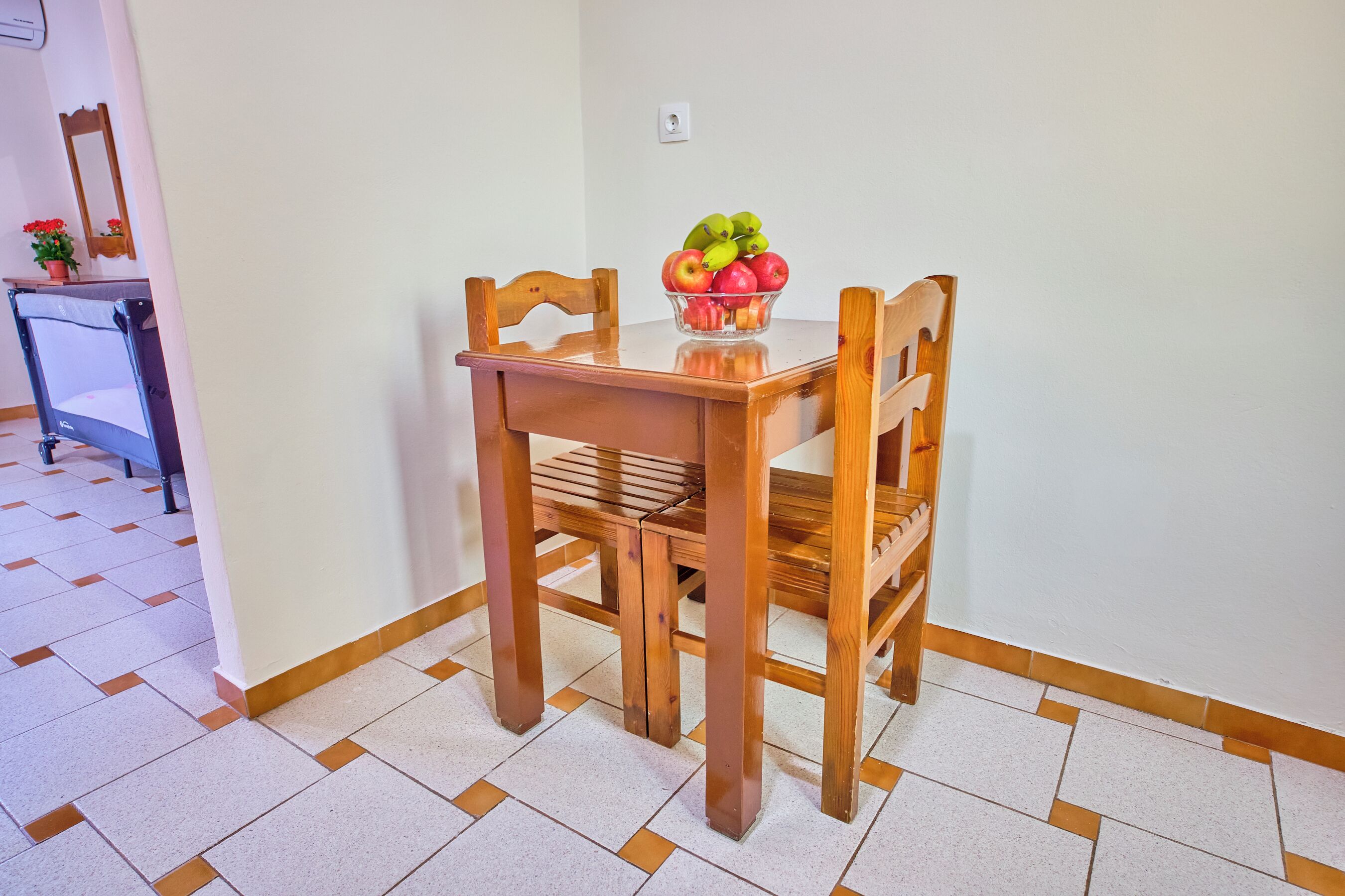 A dining table with chairs, a bowl of fruit, and a potted plant on a tiled floor.