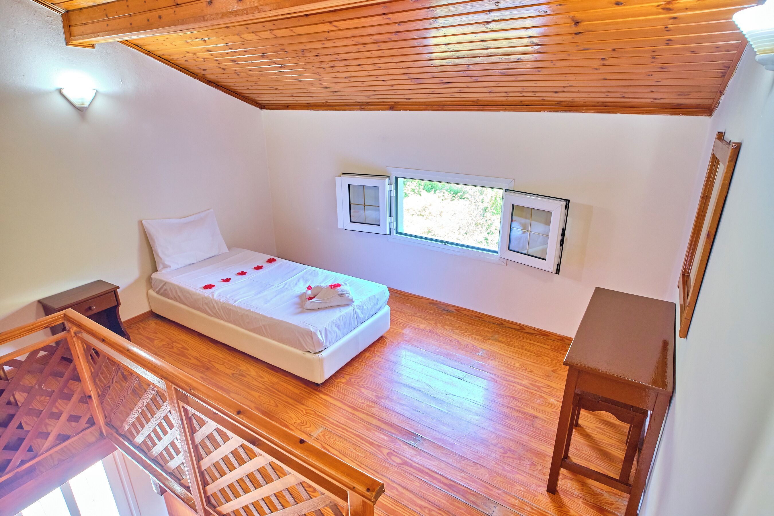 A bedroom with a wooden floor, a bed with white bedding, a nightstand, a wooden chair, and a window with a view of greenery.