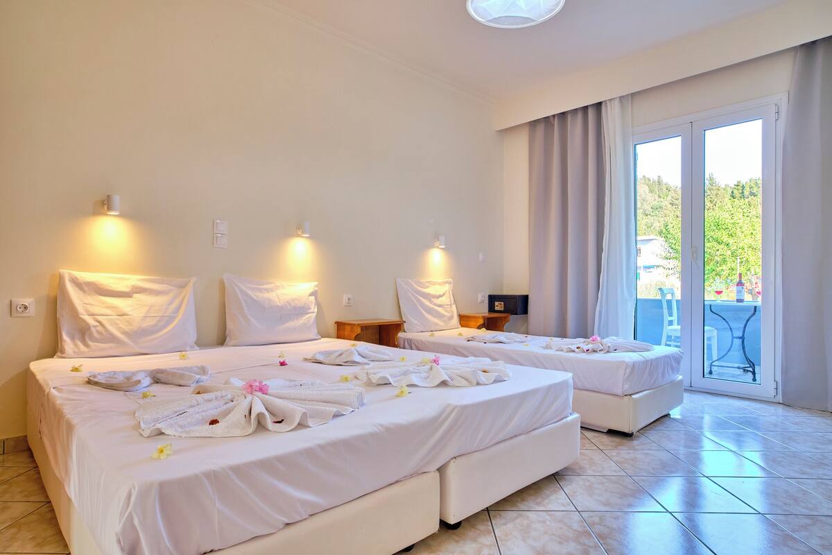 A hotel room with two beds, each with white linens and towels, a small table, and a view of a balcony with greenery outside.