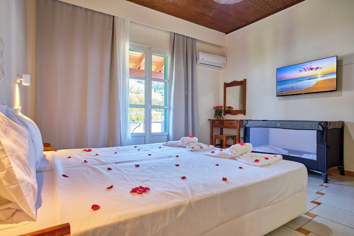 A hotel room with a large bed, a wooden headboard, a desk, a chair, an air conditioning unit, and a painting of a beach scene.