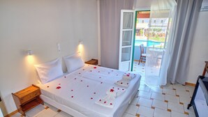 Hotelkamer met een bed, nachtkastje, een klein tafeltje met een fles en uitzicht op een zwembad en terras.