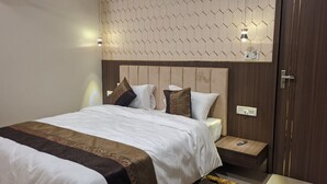 A neatly made bed with a patterned bedspread, a bedside table with a remote control, and a wall-mounted light fixture. - Shree Dhara Heaven (Varanasi)
