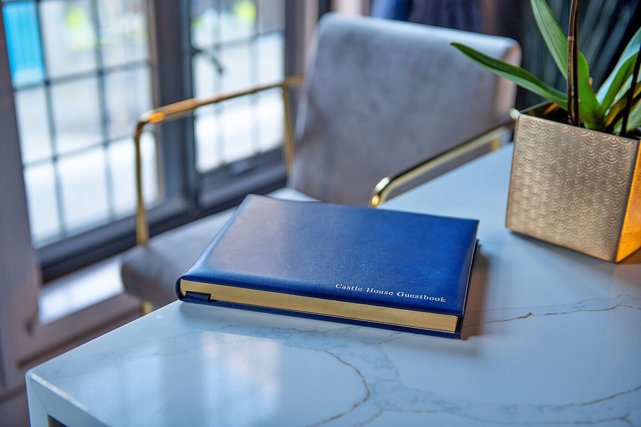 A blue book titled 'Castle House Guestbook' on a marble table next to a chair and a plant in a textured pot.