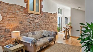 Un salón acogedor con pared de piedra, ventana con marco de madera, sofá con una manta, una mesita de noche con lámpara y una planta en maceta.