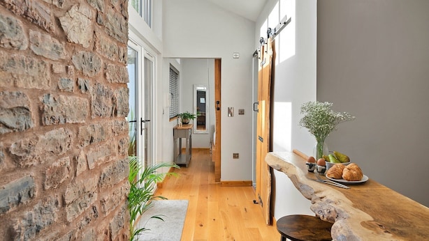 Un pasillo estrecho con suelo de madera, pared de ladrillo a un lado y un mostrador de madera con pasteles y un jarrón de flores al otro.