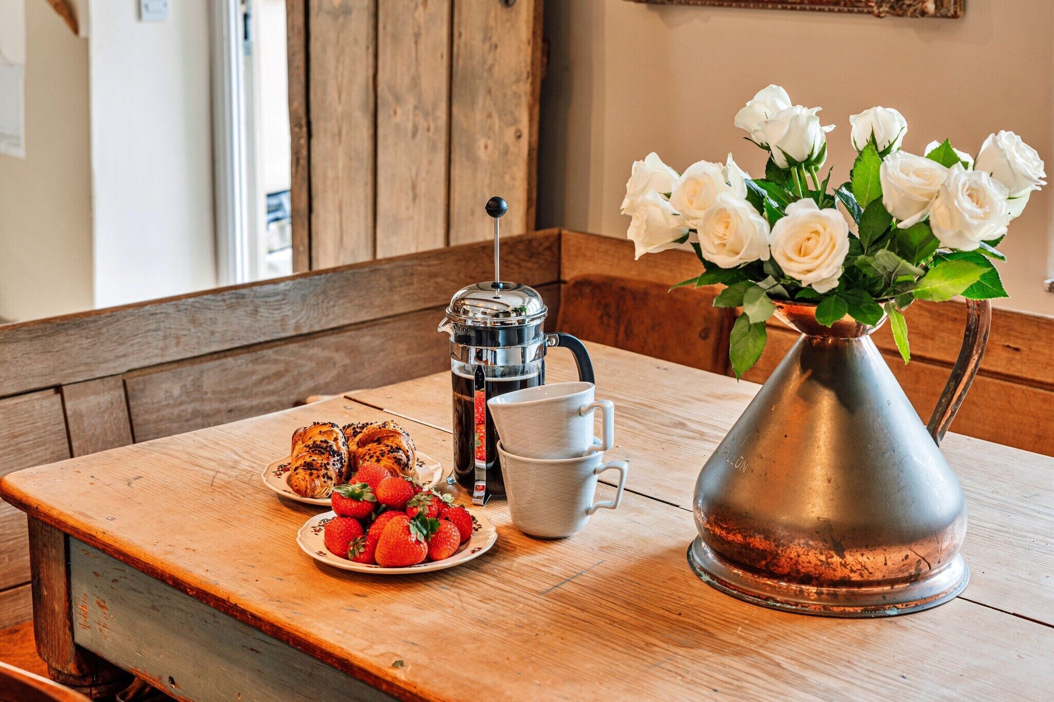 Une table en bois, une cafetière en métal, une tasse blanche, une assiette de fraises et un vase de roses blanches.
