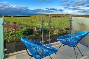 A balcony with two blue chairs, a metal railing, and a view of a green field and sea. - Chalet in Tintagel with Stunning Sea Views (Tintagel)