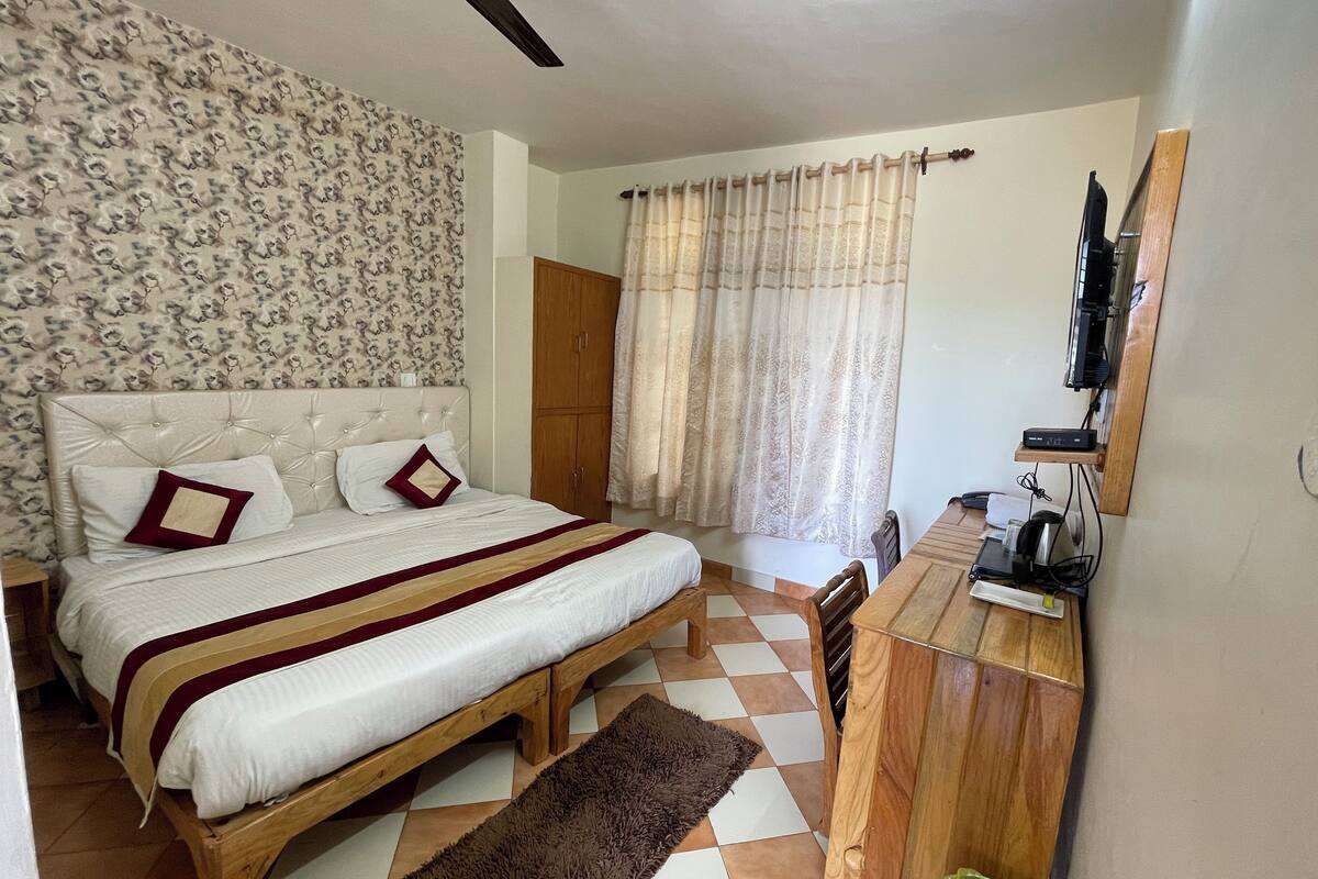 A bedroom with a bed, wooden furniture, a ceiling fan, a window with curtains, and a television on a wooden shelf.