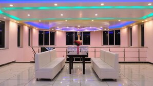 A modern hotel room with a white sofa set, a dining table with a vase of flowers, and a ceiling with colorful LED lighting. - Pink Park By Panache Hotel (Bir)