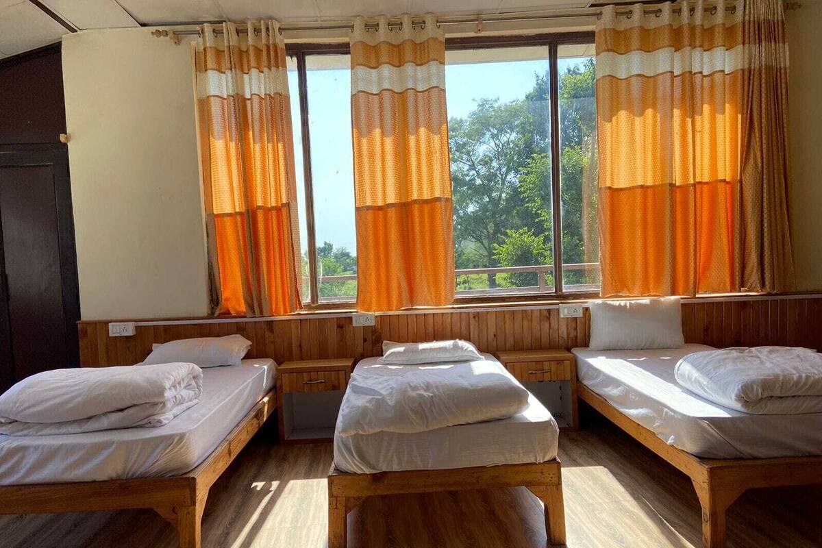 A room with three wooden beds, a window with orange curtains, and a view of greenery outside.