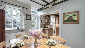 Une salle à manger avec une table en bois dressée pour quatre personnes, un vase de fleurs et une vue sur la cuisine par une porte ouverte.