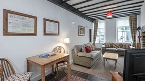 Un salon chaleureux avec une table en bois, un canapé et un téléviseur. On peut voir des certificats encadrés accrochés au mur, ainsi qu’une lampe suspendue rouge.