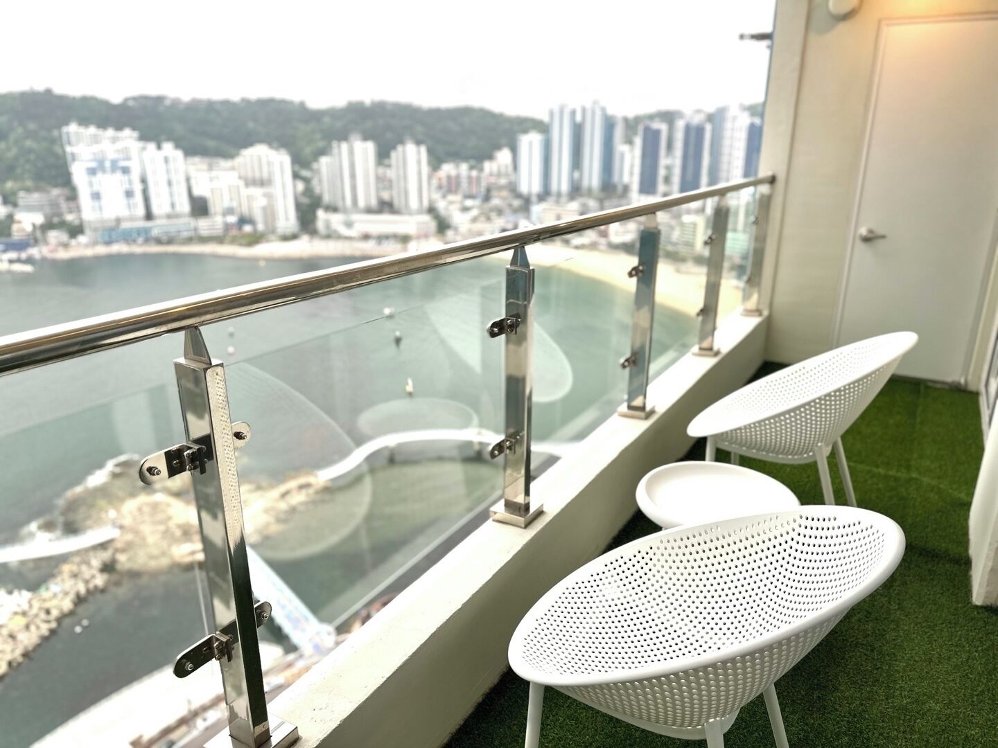 Un balcon doté d’une balustrade en verre, offrant une vue sur le paysage urbain et une étendue d’eau. On y trouve deux chaises blanches et une petite table.