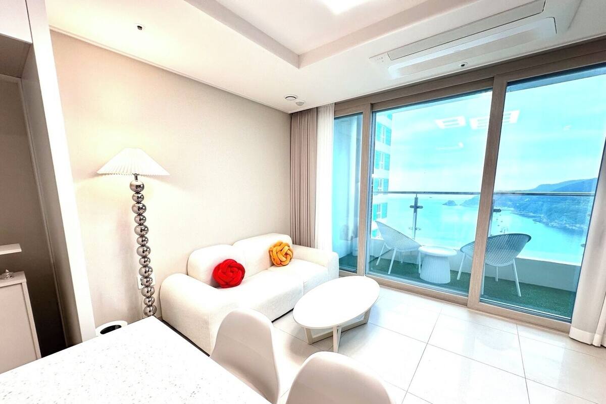 Una habitación de hotel moderna con balcón, un sofá blanco, una mesita redonda y una lámpara.