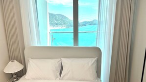 A hotel room with a bed, a nightstand, and a view of a coastal landscape through a window. - THE DECEMBER STAY Busan Ocean Terrace (Busan)