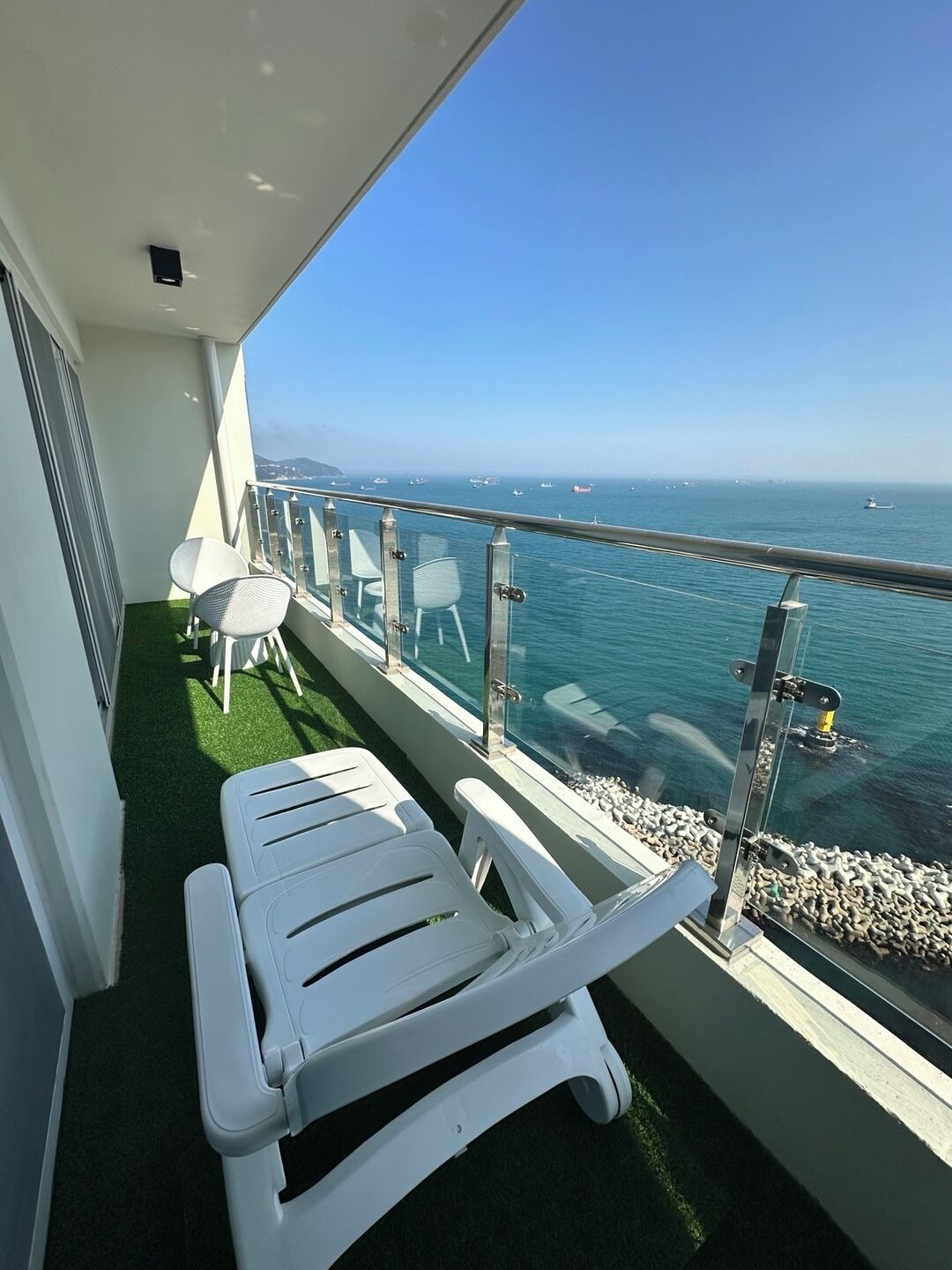 A balcony with white lounge chairs, a glass railing, and a view of the sea and boats.