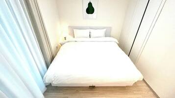 Una cama bien hecha en una habitación con piso de color claro y una única lámpara de pared.