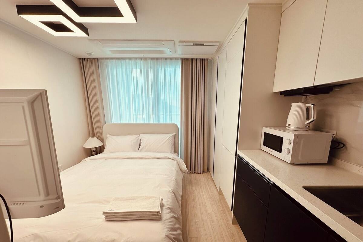 Habitación de hotel con cama, mesita de noche con lámpara, microondas y una pequeña cocina.