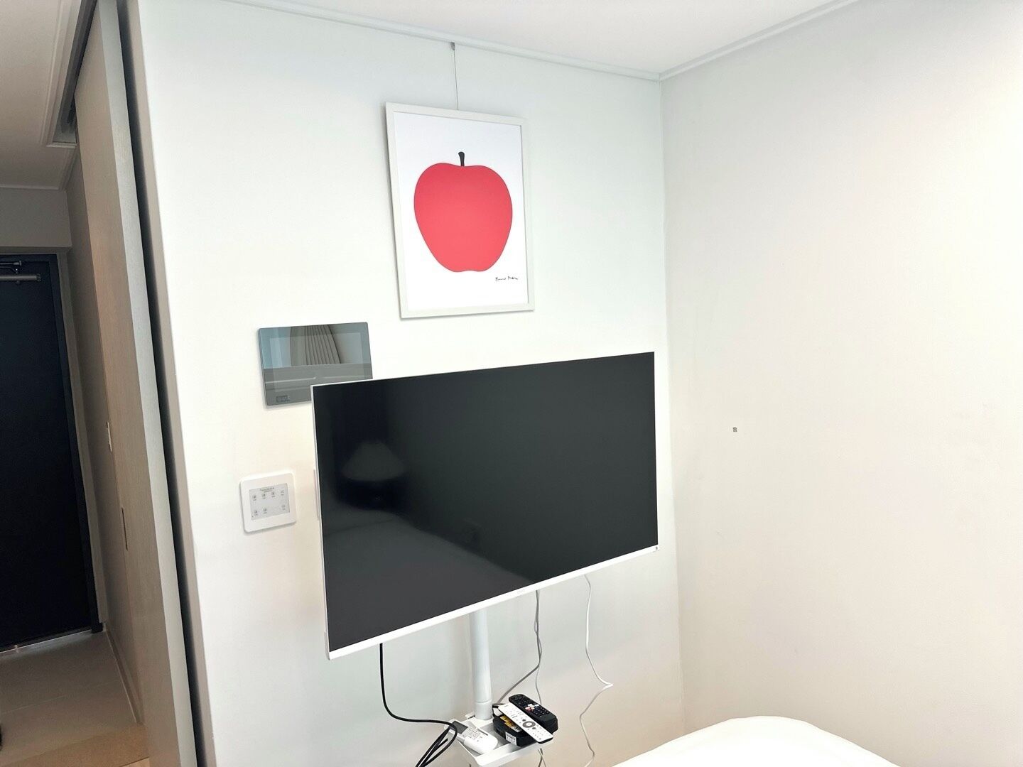 A modern room with a large flat-screen TV mounted on the wall, a framed picture of an apple above it, and a small control panel to the left.
