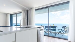 A modern kitchen with a dishwasher, a dining area with a table and chairs, and a large sliding glass door leading to a balcony with a view of the sea. - Mar Apartment by Atlantic Holiday (Camara de Lobos)