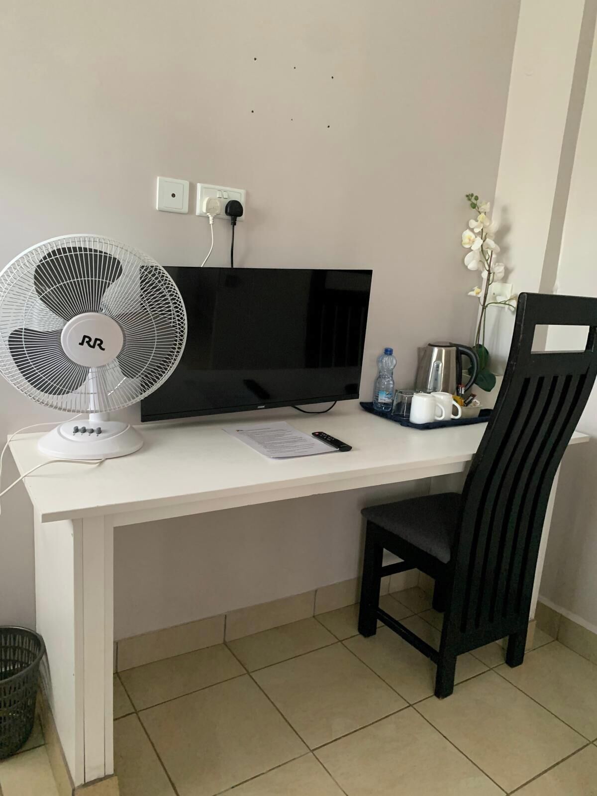 A white desk with a black chair, a black TV, a white fan, a remote control, a water bottle, a coffee maker, and a small plant.
