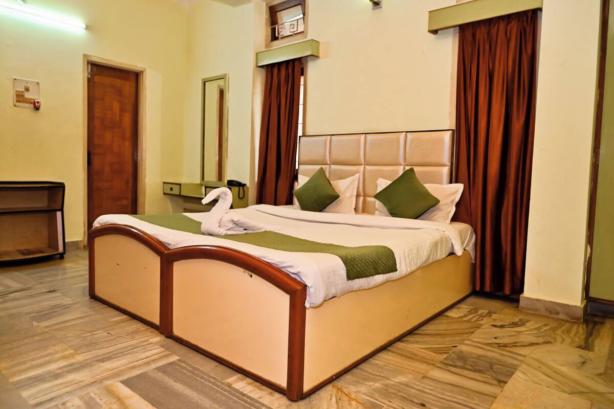 A hotel room with a bed, a wooden headboard, a green throw blanket, and brown curtains.