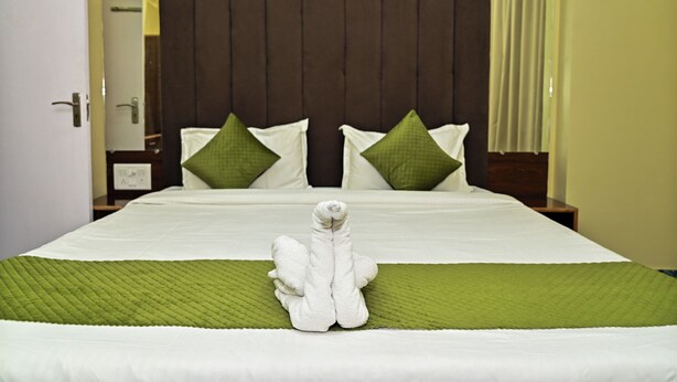 A neatly made bed with a green bedspread and pillows, a white towel folded on the bed, and a dark headboard. - Holiday Inn Guest House (Guwahati)