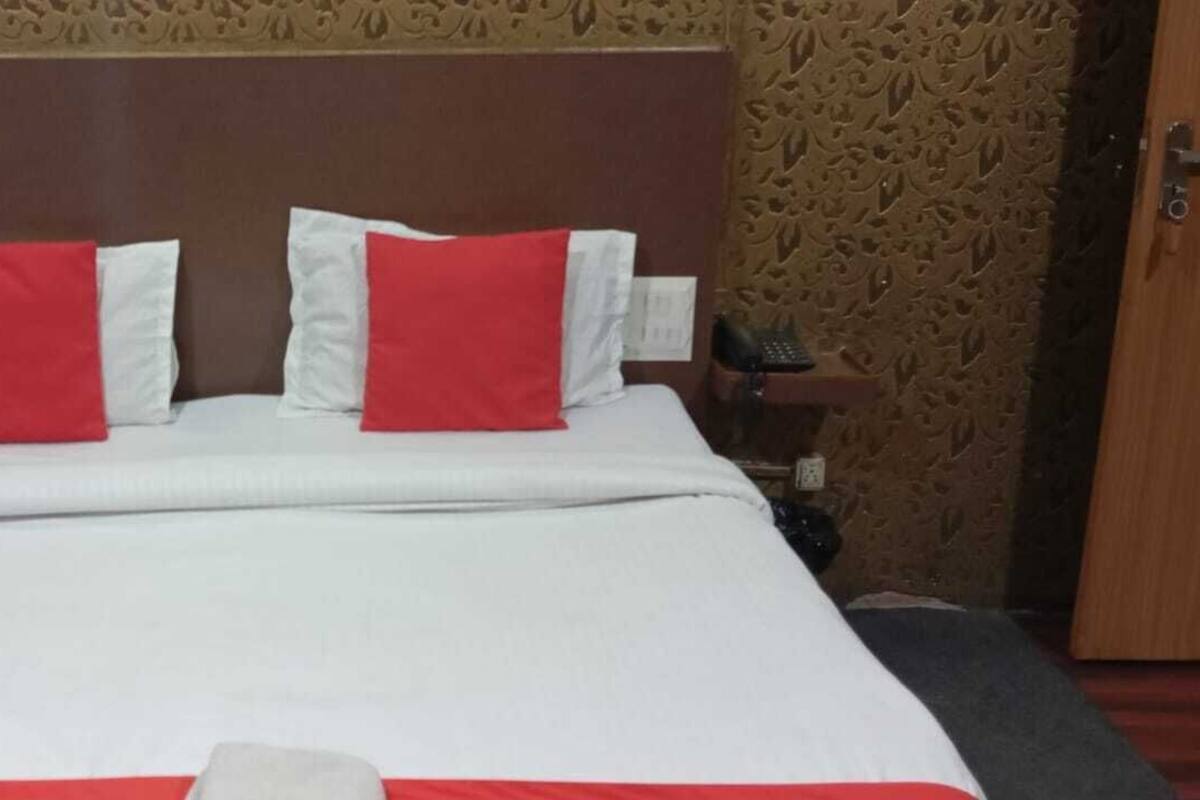 A neatly made bed with white bedding and two red pillows, a wooden headboard, and a patterned wall in the background.