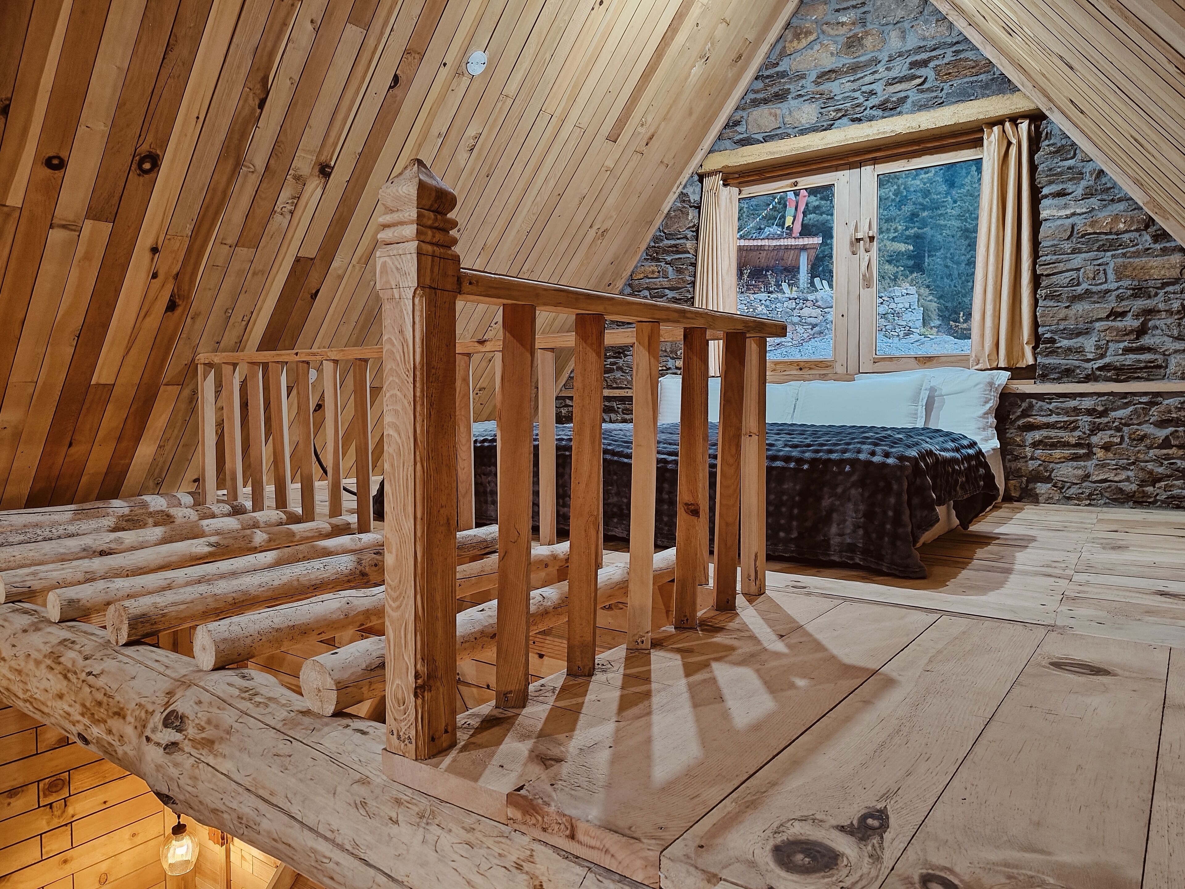 A wooden staircase leading to a loft bedroom with a stone wall and a window overlooking a snowy landscape.
