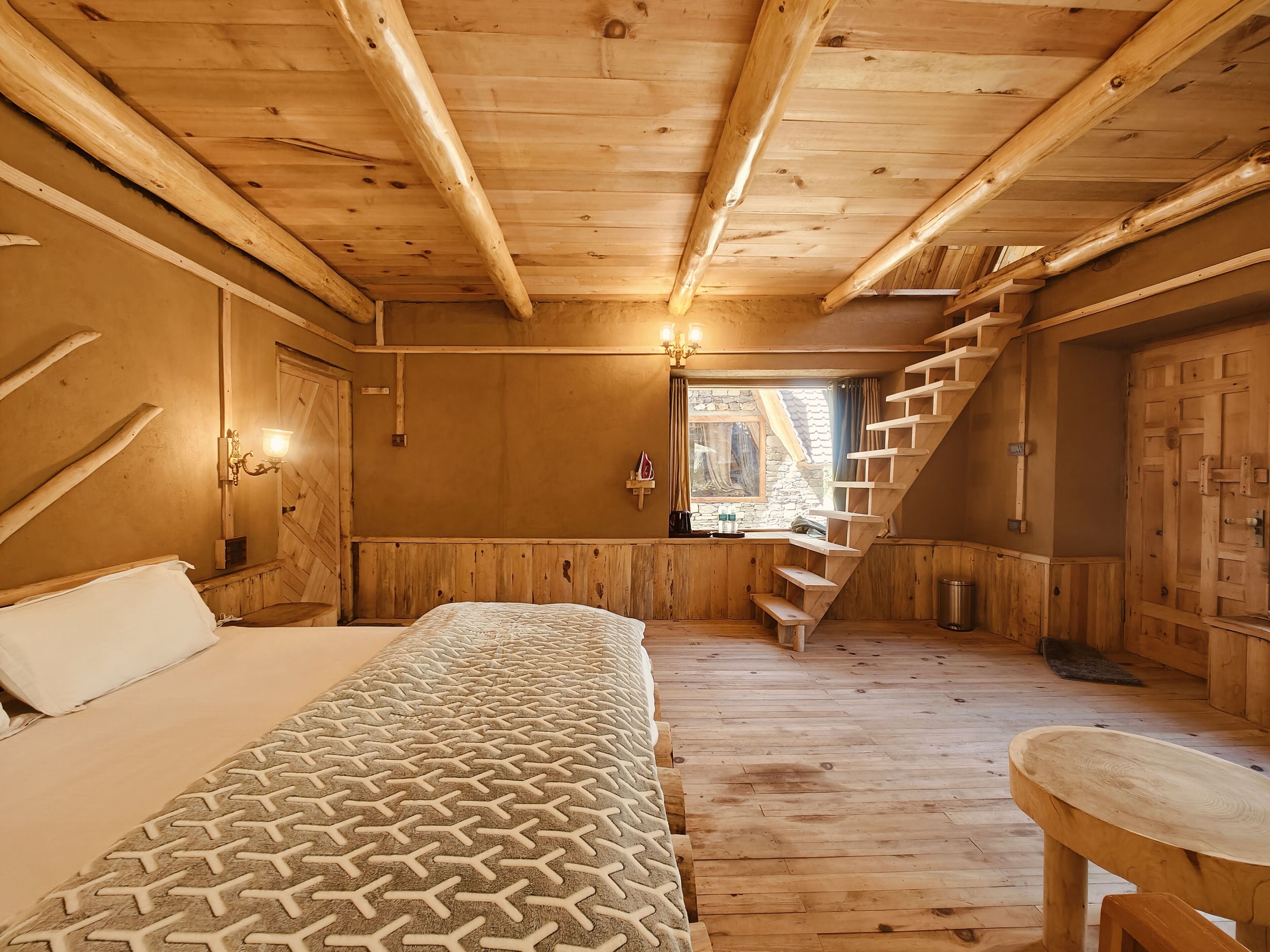 A wooden cabin room with a bed, a small table, a staircase, and a window.
